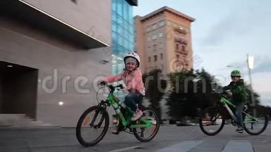 <strong>一个</strong>微笑的<strong>小</strong>女孩和男孩骑绿色自行车在<strong>一个</strong>城市的晚上在<strong>一个</strong>圆圈。 戴着头盔的<strong>小</strong>自行车